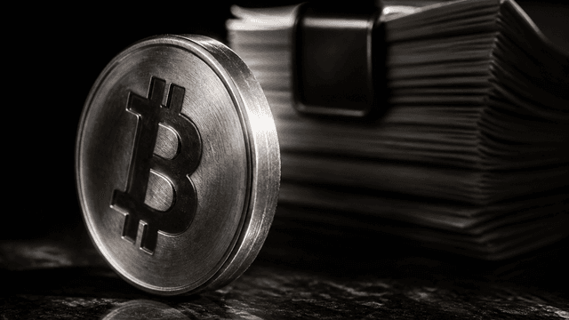 Metallmünze mit eingeprägtem Bitcoin-Zeichen steht hochkant vor einem Stapel gebundener Antragsdokumente