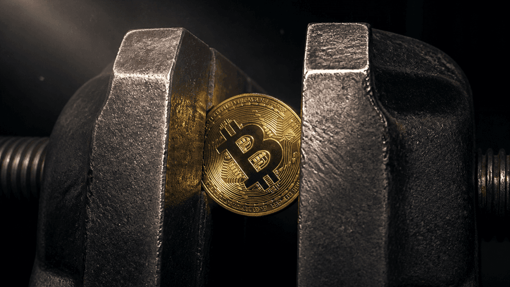 Goldene Münze mit Bitcoin-Symbol verkeilt zwischen den Stahlbacken eines Schraubstocks vor einem dunklen Metallkanal