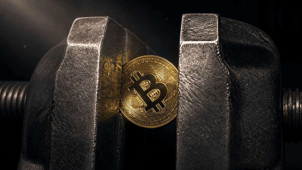 Goldene Münze mit Bitcoin-Symbol verkeilt zwischen den Stahlbacken eines Schraubstocks vor einem dunklen Metallkanal