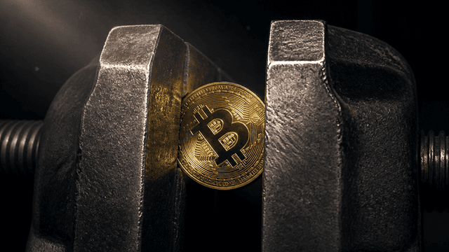 Goldene Münze mit Bitcoin-Symbol verkeilt zwischen den Stahlbacken eines Schraubstocks vor einem dunklen Metallkanal