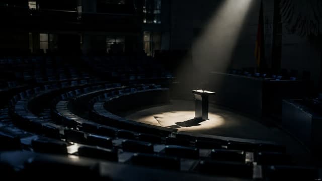 Leerer Plenarsaal eines Parlaments bei Nacht, ein harter Lichtkegel auf dem Rednerpult