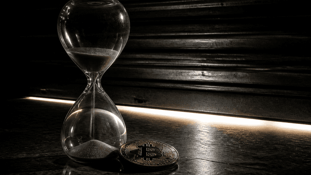 Fast leergelaufene Sanduhr neben einer Muenze mit Bitcoin-Zeichen vor einem herabgefahrenen Rolltor