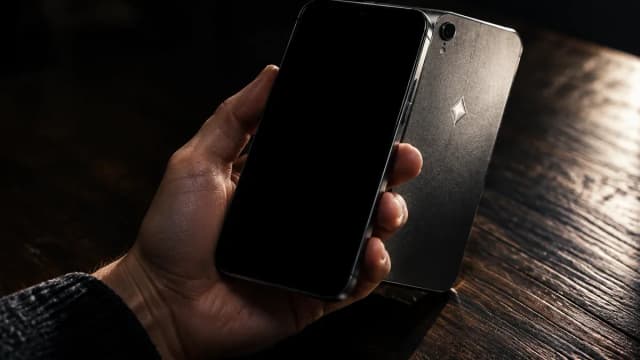 Eine Hand hält ein markenloses Smartphone mit dunklem Display über einem Holztisch, auf der metallenen Rückseite blitzt ein eingraviertes rautenförmiges Symbol auf.