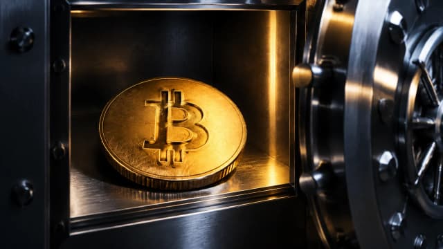 Eine goldene Münze mit dem Bitcoin-Zeichen liegt in einem geöffneten Schließfach aus gebürstetem Stahl, daneben die halb offene Tresortür