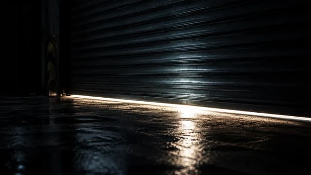 Schweres Stahl-Rolltor einer Lagerhalle faehrt herunter und steht nur noch einen Lichtspalt offen ueber nassem Betonboden