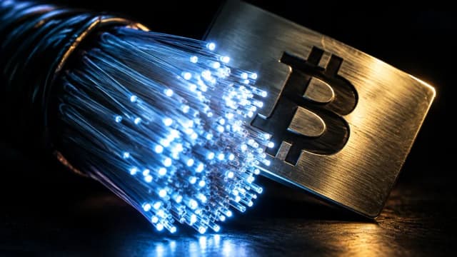 Makro eines Bündels Glasfaserkabel, dessen Enden im Dunkeln hell aufleuchten, daneben eine Messingplakette mit eingraviertem Bitcoin-Zeichen