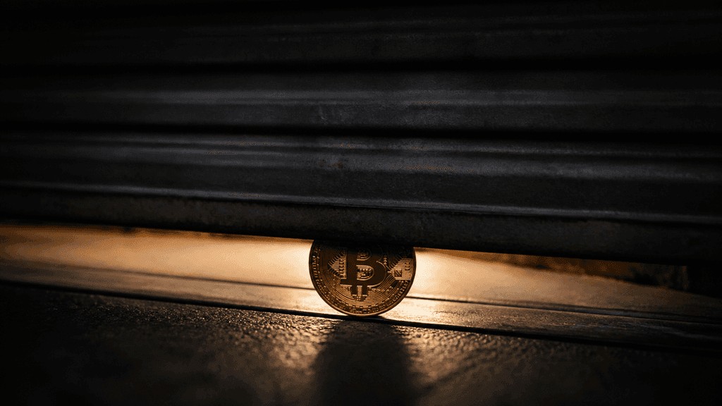 Ein herabgelassenes Metall-Rolltor lässt nur noch einen schmalen Lichtspalt offen, im Spalt steht aufrecht eine goldene Münze mit eingeprägtem Bitcoin-Symbol.