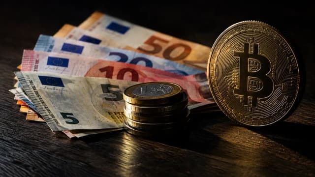 Aufgefächerte Euro-Geldscheine und ein Stapel Euro-Münzen neben einer aufrecht stehenden Metallmünze mit eingeprägtem Bitcoin-Symbol auf dunklem Holz
