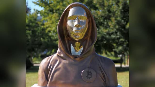 Bronzebüste von Satoshi Nakamoto mit Kapuze, spiegelndem Gesicht und Bitcoin-Zeichen auf der Brust im Graphisoft Park in Budapest